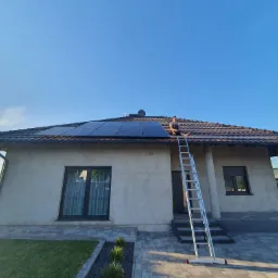 Instalator siedzi na drabinie opierającej się o dach domu z panelami słonecznymi, błękitne niebo w tle.