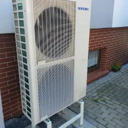 Biała pompa ciepła Samsung na stalowej konstrukcji, zamontowana przy ścianie budynku z czerwoną cegłą i szarą elewacją, widoczna mała doniczka z czerwonymi kwiatami w oknie.