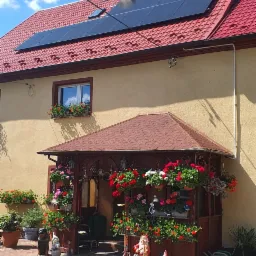 Żółty dom jednorodzinny z czerwonym dachem i panelami fotowoltaicznymi na dachu, ozdobiony kwiatami w oknach i donicach, oraz licznymi figurkami gnomów w ogrodzie.