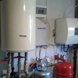 Energy Power Group Sp. Z o.o - Biały zbiornik ciepłej wody użytkowej z podłączoną instalacją hydrauliczną, srebrny i czerwony zbiornik wyrównawczy ciśnienia, skrzynka elektryczna Samsung na białej ścianie.