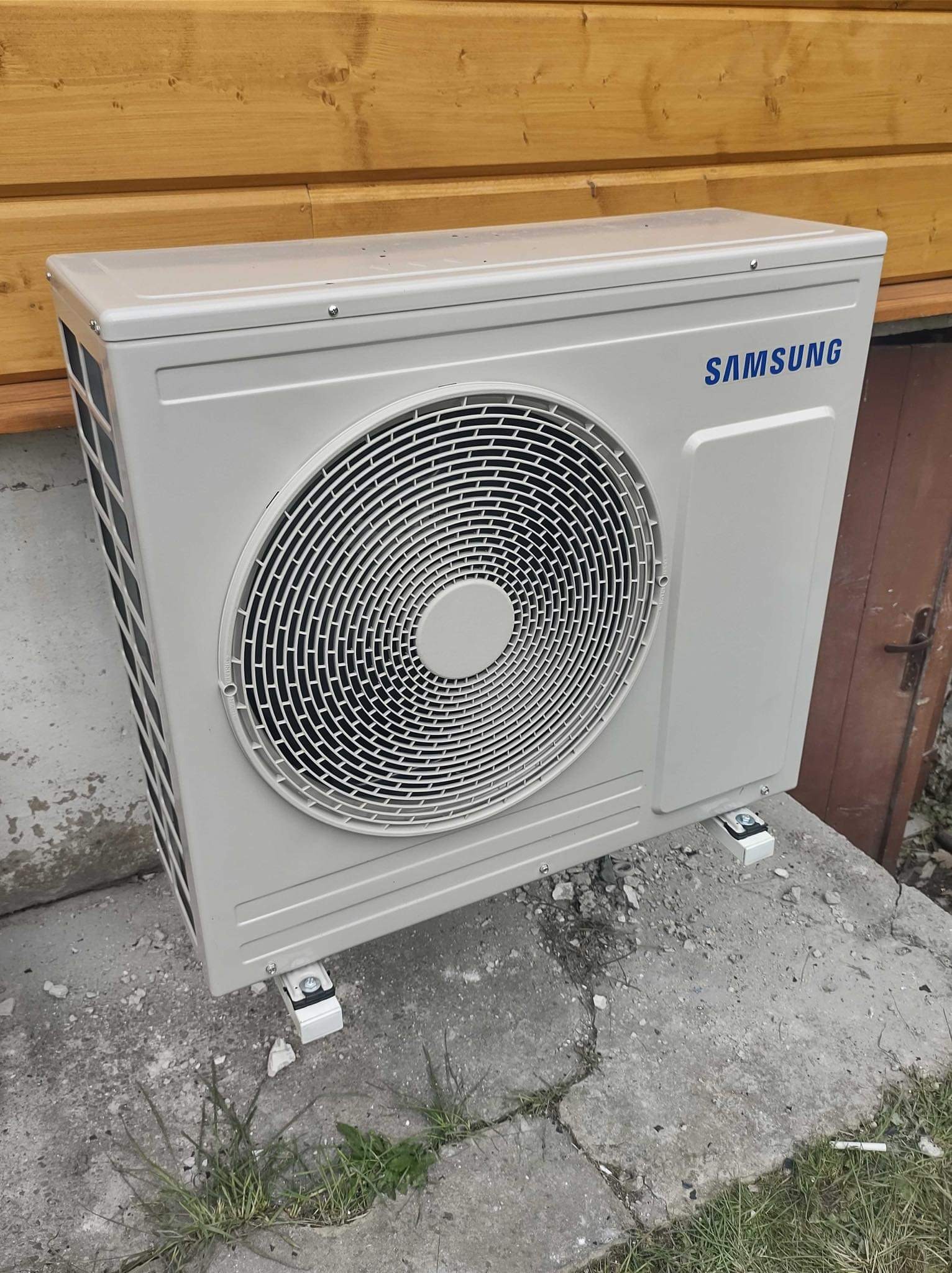 Zewnętrzna jednostka klimatyzacyjna marki Samsung, zamontowana na betonowym fundamencie, z drewnianą ścianą w tle.