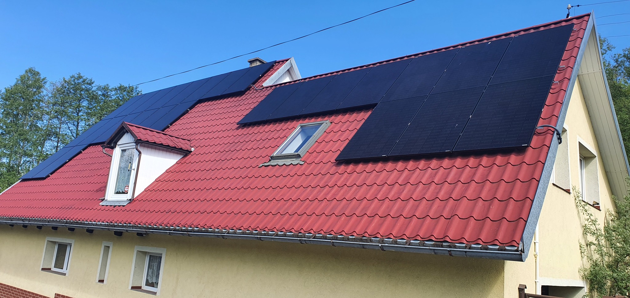 Czerwony dach dwuspadowy z oknami dachowymi i panelami fotowoltaicznymi w słoneczny dzień.