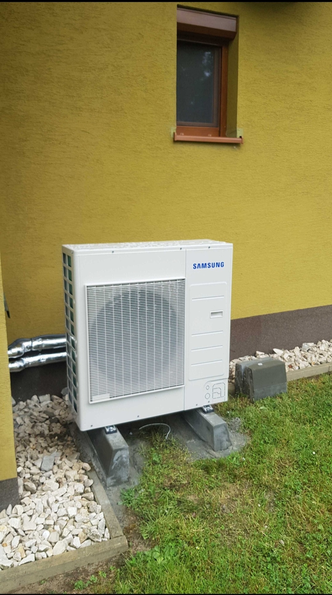 Biała zewnętrzna jednostka klimatyzacyjna Samsung na betonowych wspornikach, ustawiona przy żółtej ścianie budynku z małym oknem, obok rury i kamieni.