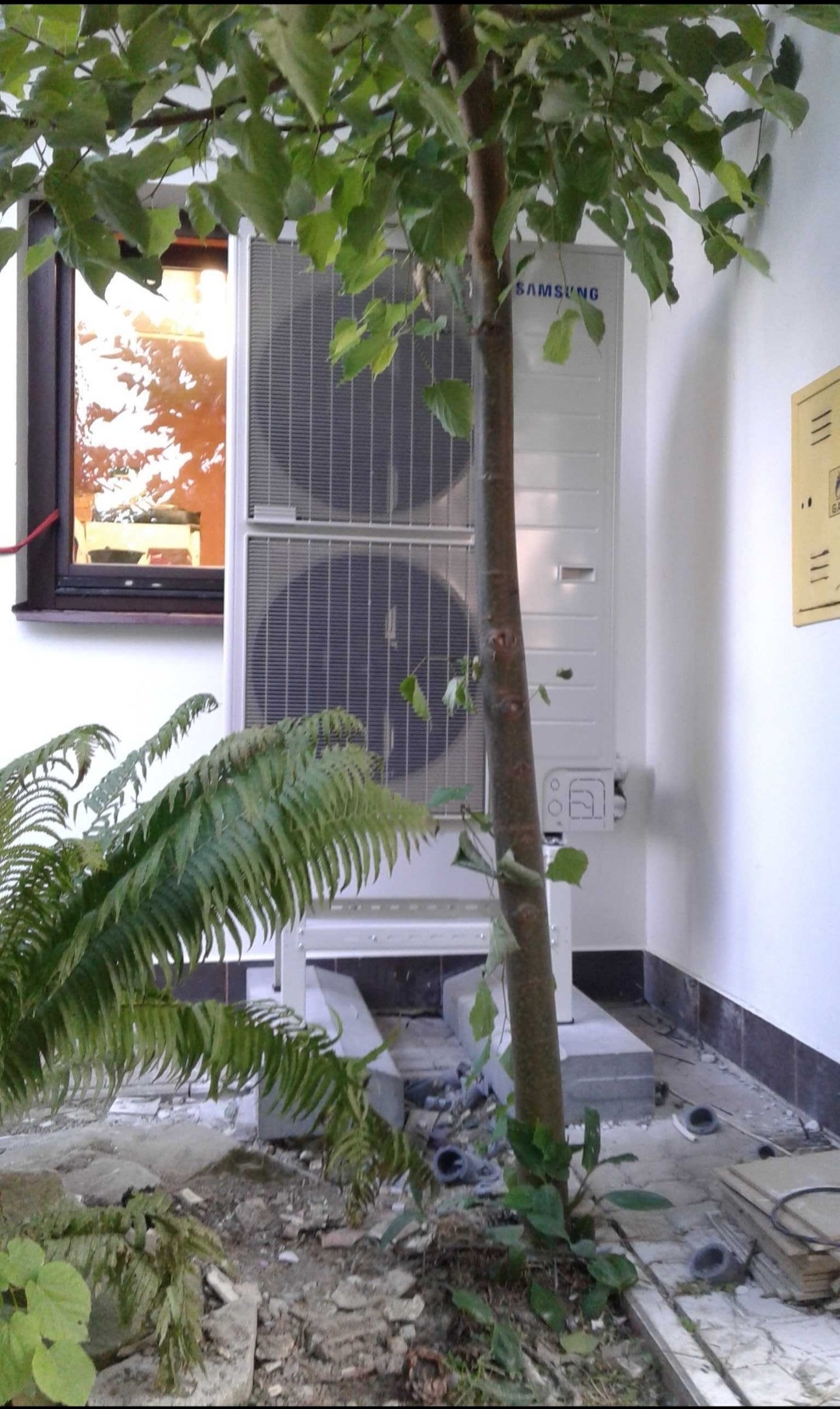 Biała zewnętrzna jednostka klimatyzacji Samsung, częściowo zasłonięta przez pień drzewa, z widocznym oknem i paprocią w tle, na zewnątrz budynku.