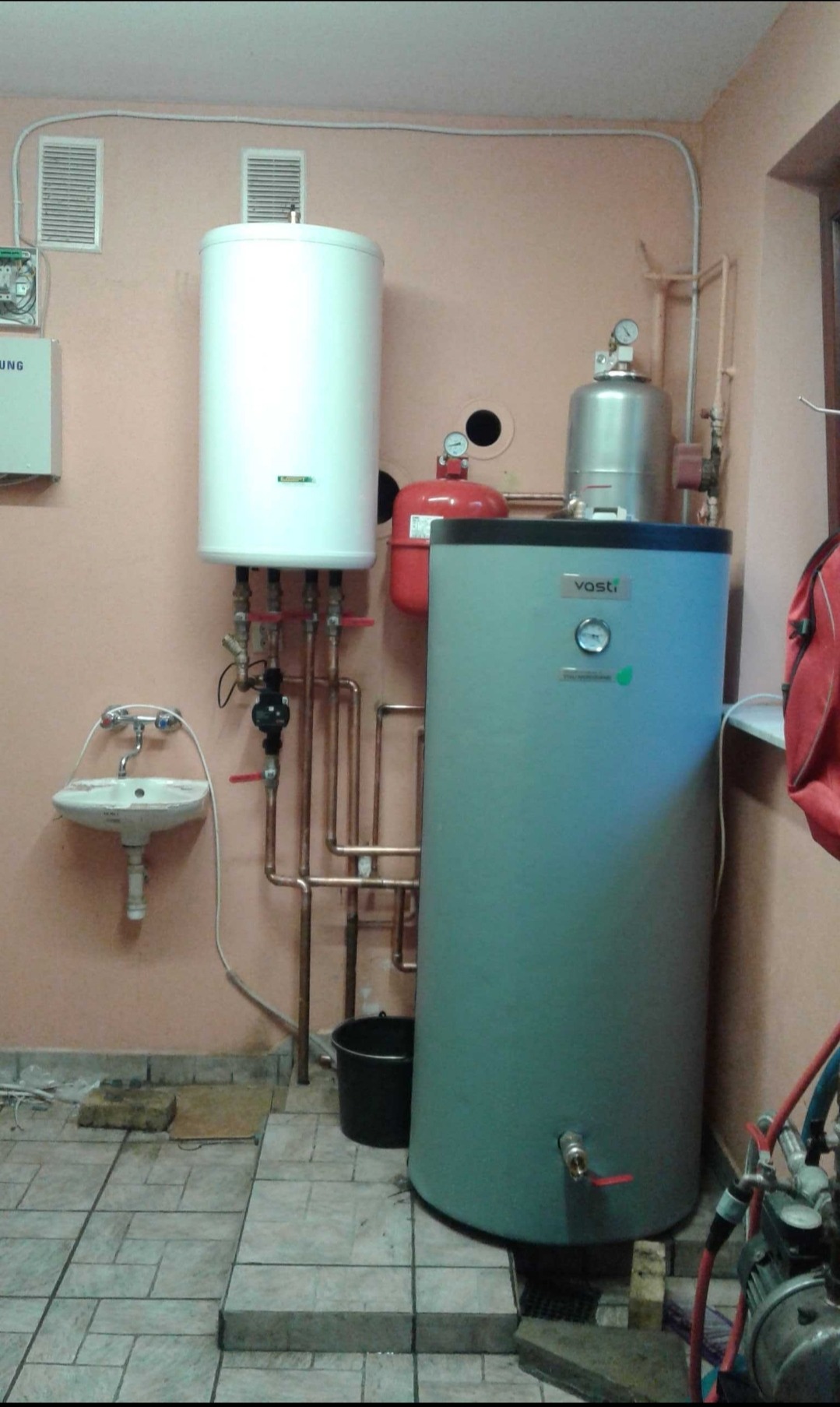 Instalacja dwóch zbiorników buforowych z armaturą w pomieszczeniu kotłowni, widoczna umywalka i fragment sprężarki.