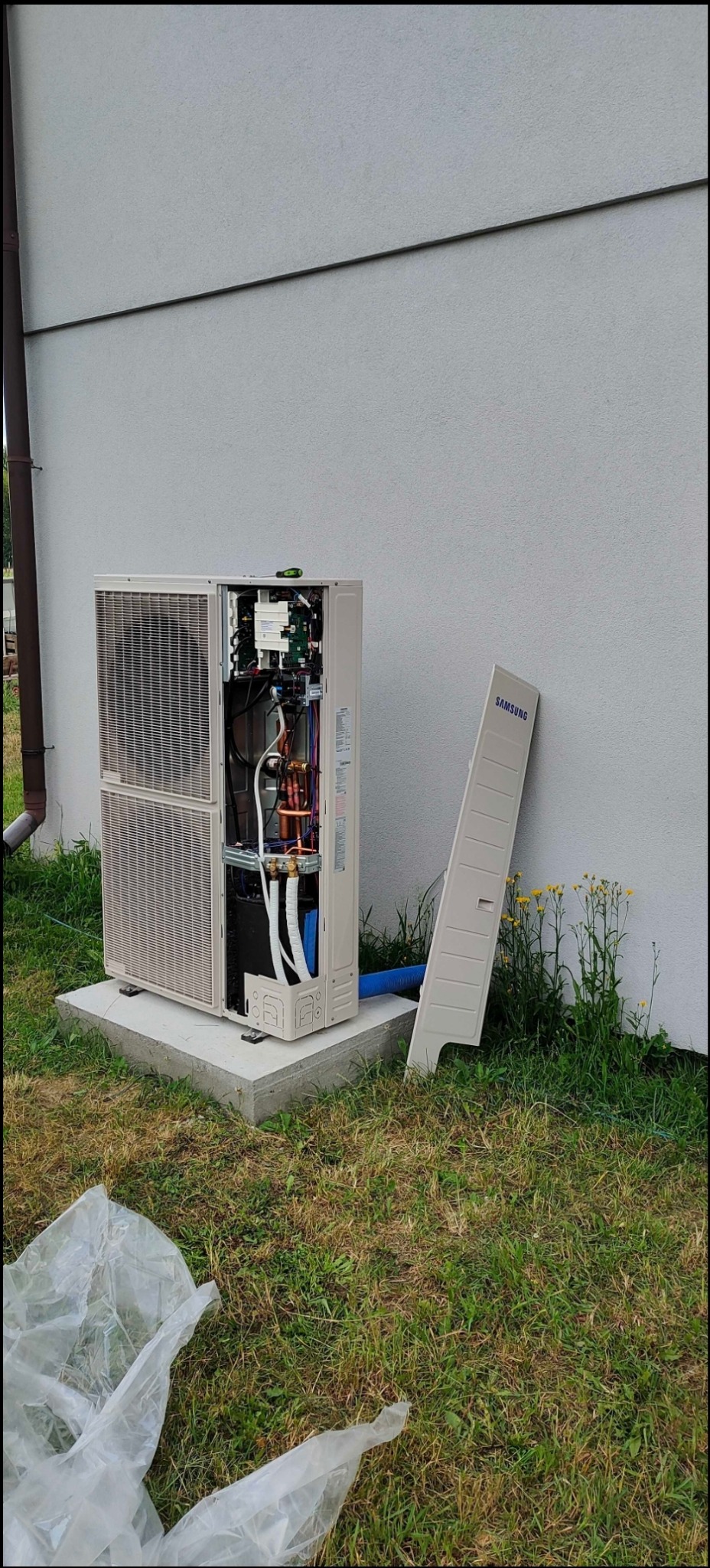 Otwarta obudowa zewnętrznej jednostki klimatyzacyjnej Samsung na betonowej podstawie, z widocznymi miedzianymi rurami i przewodami, obok ściany budynku i żółtych kwiatów, zdemontowany panel oparty...