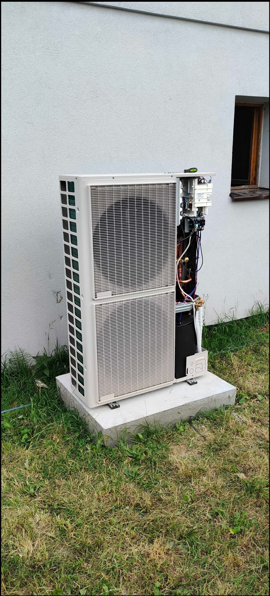 Biała jednostka zewnętrzna z otwartym panelem bocznym, ukazującym wewnętrzne okablowanie i komponenty; umieszczona na betonowej podstawie przy ścianie budynku z oknem.