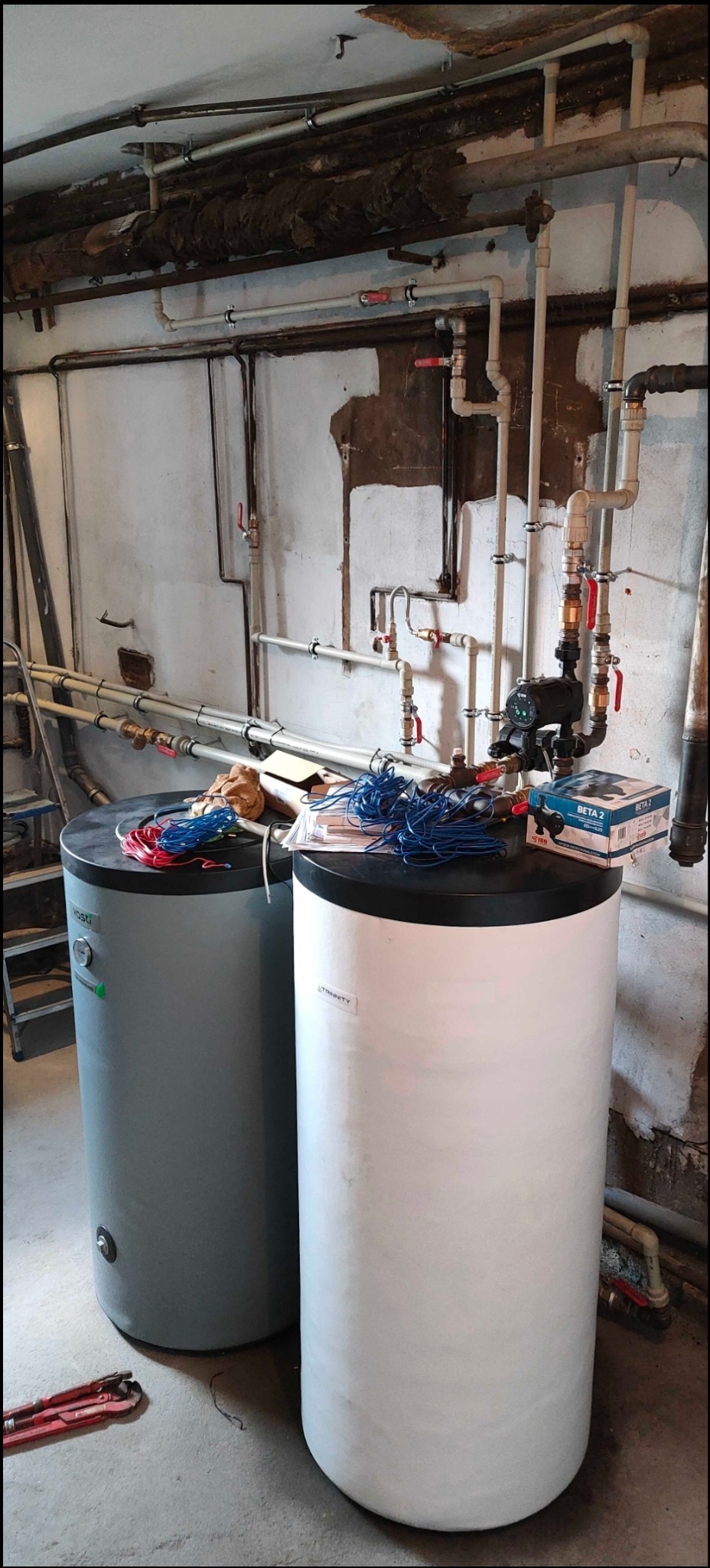 Dwa zbiorniki buforowe ciepła, szary i biały, w piwnicy w Jeleniej Górze, z widocznymi rurami i instalacją hydrauliczną na ścianie. Na zbiornikach leżą przewody elektryczne i pudełko z pompą...