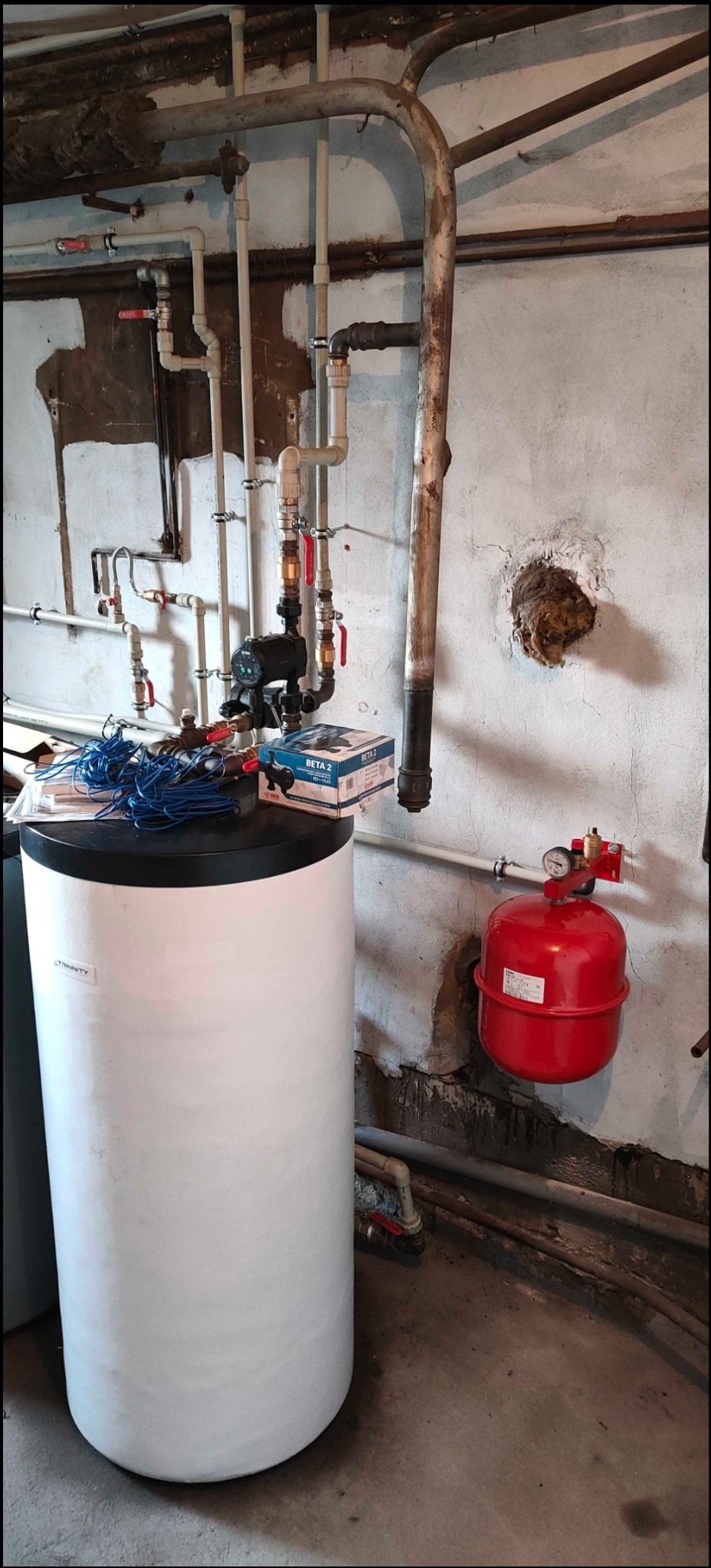 Biały zasobnik ciepłej wody użytkowej z podłączonymi rurami i grupą pompową, stojący na betonowej podłodze w piwnicy, z widocznym czerwonym naczyniem przeponowym i przewodami elektrycznymi na górze.