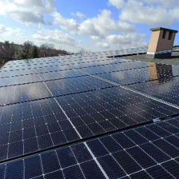 Rząd paneli słonecznych na dachu budynku, odbijających błękitne niebo z chmurami; w tle kominy wentylacyjne i drzewa.