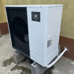 Zewnętrzna jednostka klimatyzacji Mitsubishi Zubadan umieszczona na betonowych blokach, z widoczną rurą w izolacji, na tle otynkowanej ściany.