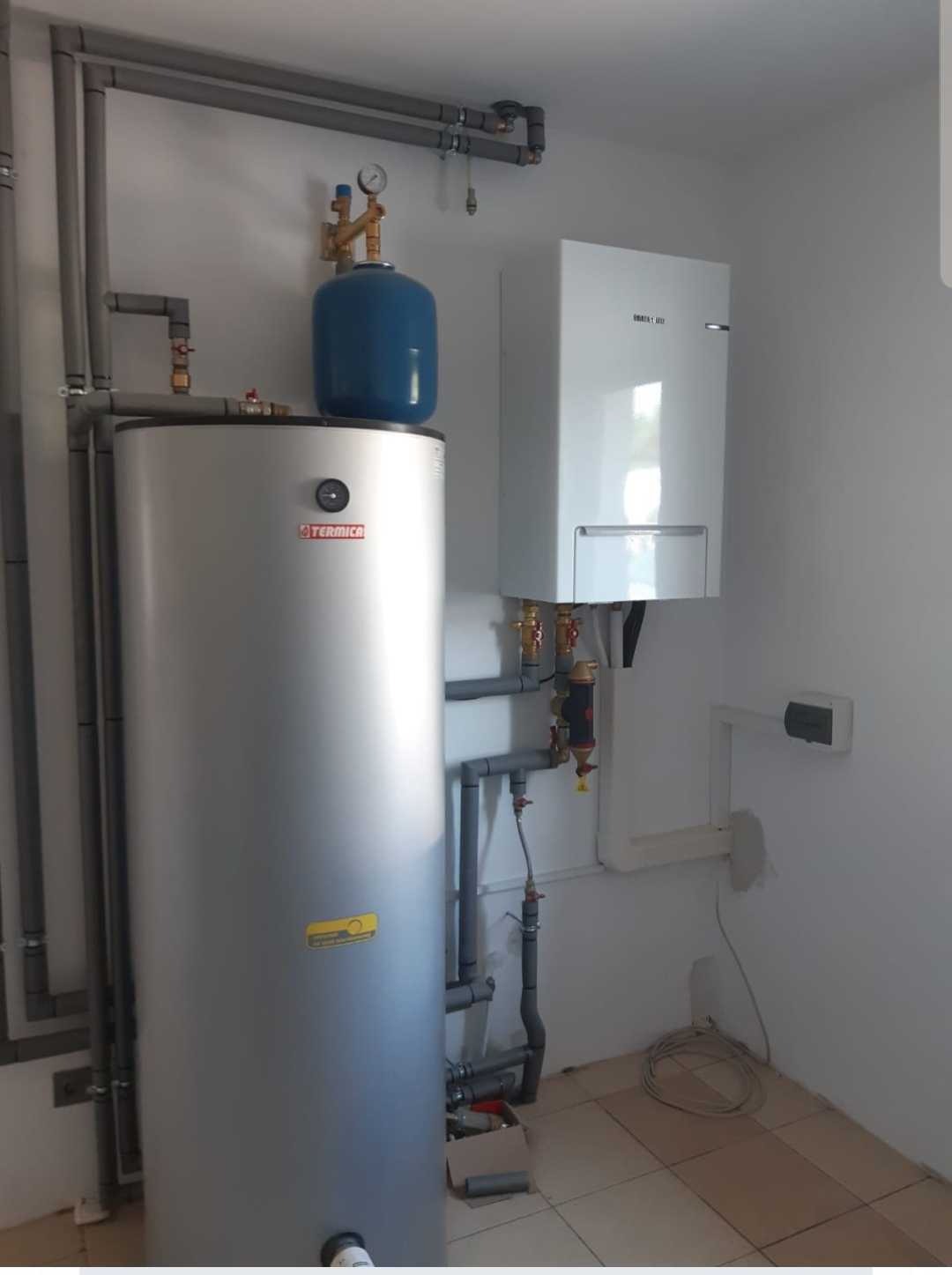 Szary, pionowy zbiornik z niebieskim zbiornikiem wyrównawczym na górze, połączony rurami z białym kotłem wiszącym na ścianie. Widoczne liczne rury i zawory.