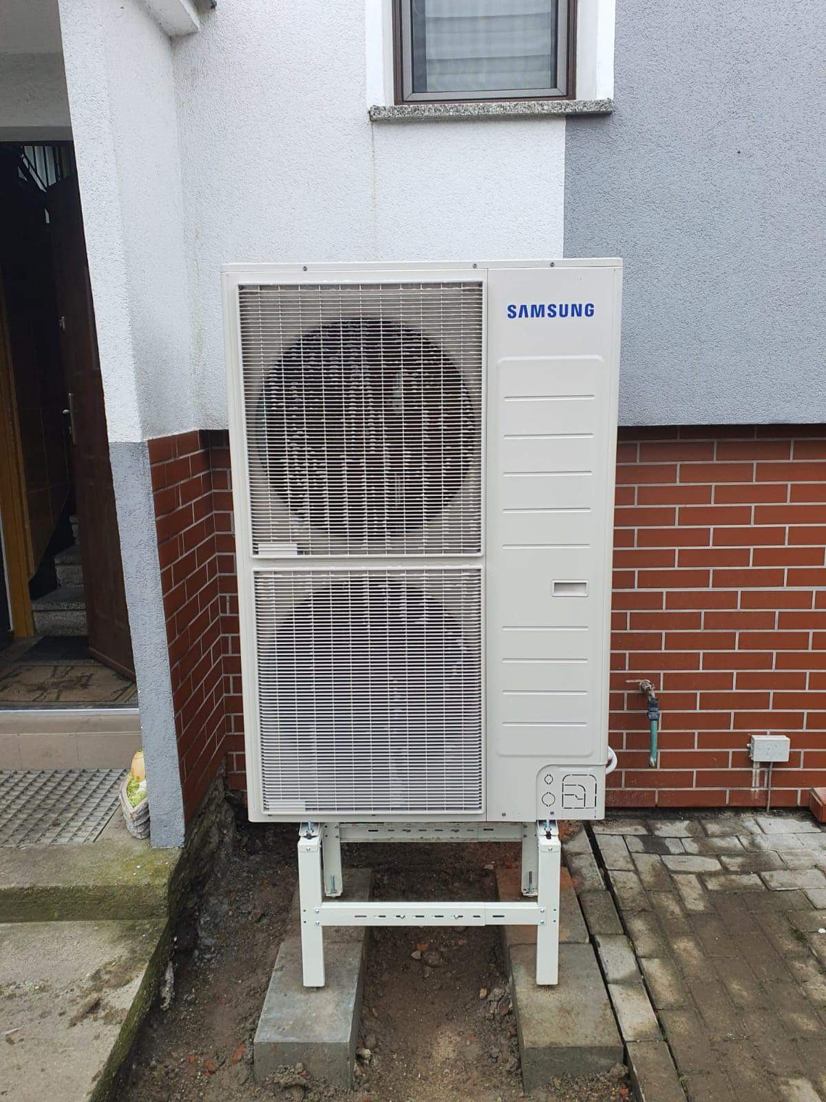 Biała zewnętrzna jednostka pompy ciepła Samsung zamontowana na wspornikach na tle ściany z cegły i szarego tynku.