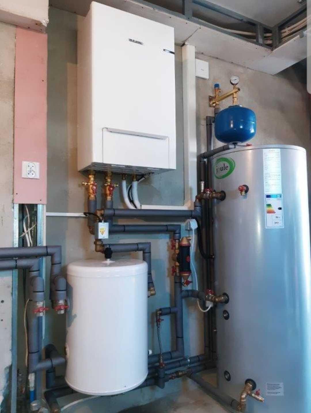 Zamontowany system grzewczy z białym kotłem wiszącym, szarymi rurami i dwoma zbiornikami: białym mniejszym i srebrnym większym z niebieskim naczyniem wzbiorczym, w surowym pomieszczeniu z widoczną...