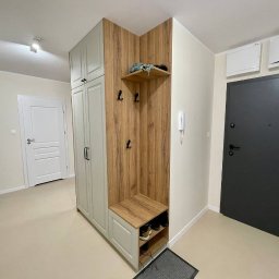 Maiya Harelik Haros.home - Funkcjonalna zabudowa przedpokoju z szafą, wieszakami i siedziskiem z półkami na buty, utrzymana w kolorystyce drewna i jasnej szarości, dopasowana do niewielkiej przestrzeni.