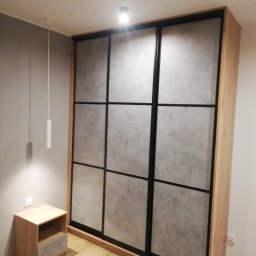 Maiya Harelik Haros.home - Duża szafa przesuwna z drewnianą ramą i czarnymi profilami, wypełniona panelami imitującymi beton, stojąca w sypialni obok drewnianej szafki nocnej z szufladą.