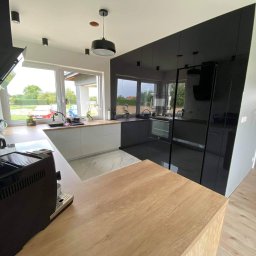 Maiya Harelik Haros.home - Nowoczesna kuchnia z czarnymi, lakierowanymi frontami szafek i białymi elementami, drewniany blat wyspy kuchennej na pierwszym planie, widok na okno z zielenią na zewnątrz.