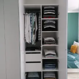 Wnętrze białej szafy na wymiar w Smolcu z drzwiami uchylnymi, widoczne ubrania na wieszakach i półkach, odbicie pokoju w lustrzanych drzwiach.