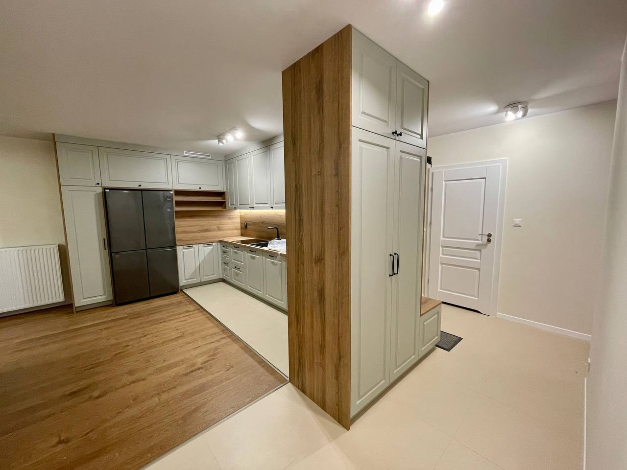 Jasnoszara zabudowa kuchenna z drewnianym blatem i panelem ściennym, połączona z szafą w przedpokoju, tworząca spójną całość w nowoczesnym wnętrzu.