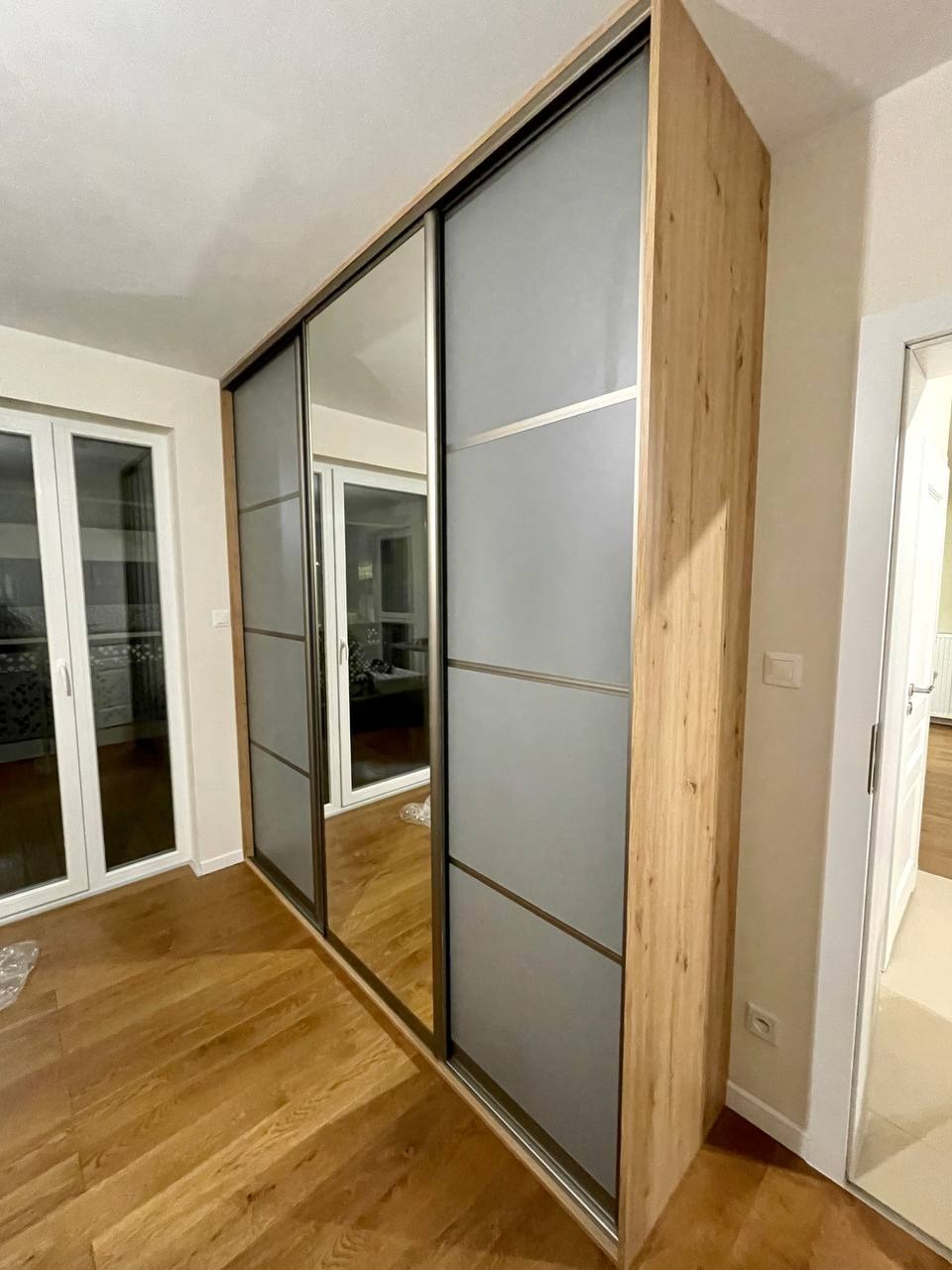 Szafa wnękowa z przesuwnymi drzwiami, fronty z lustrami i matowego szkła, obudowa z płyty drewnopodobnej, parkiet na podłodze.