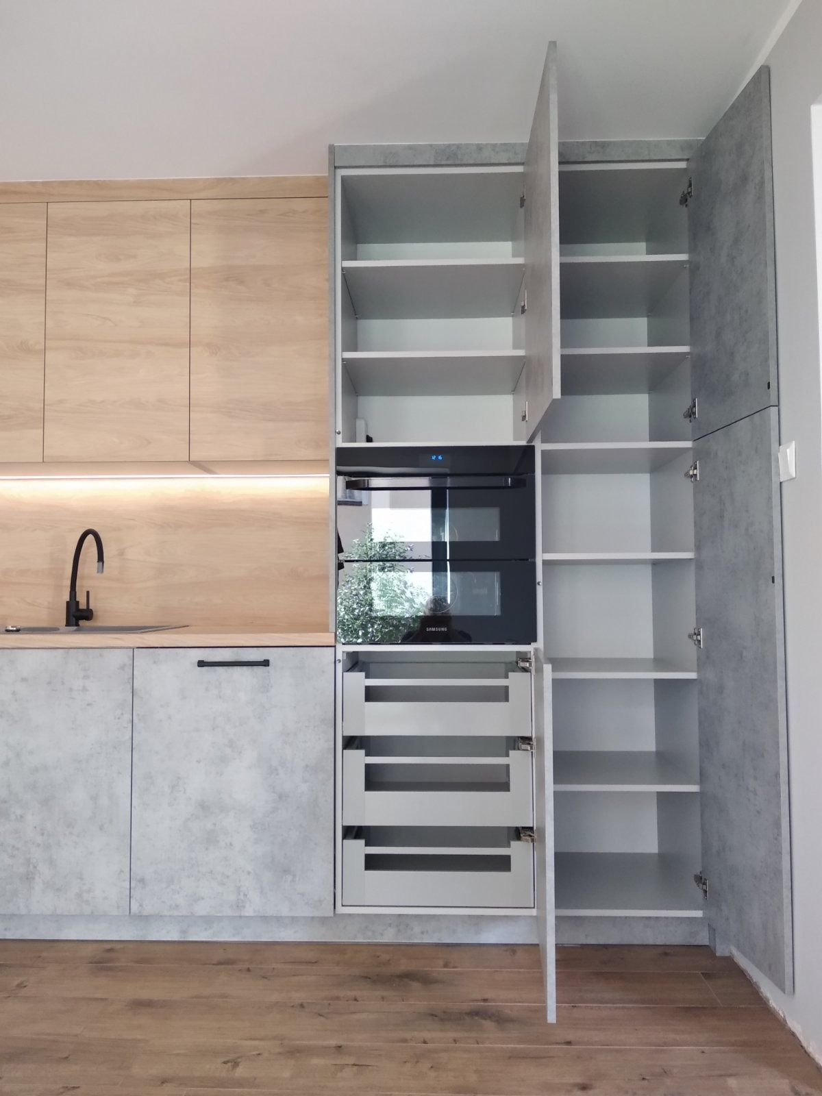 Nowoczesna zabudowa kuchenna z frontami imitującymi beton, drewnianymi szafkami i czarną armaturą, wnęka z piekarnikiem i wysuwanymi półkami, otwarte szafki z półkami.