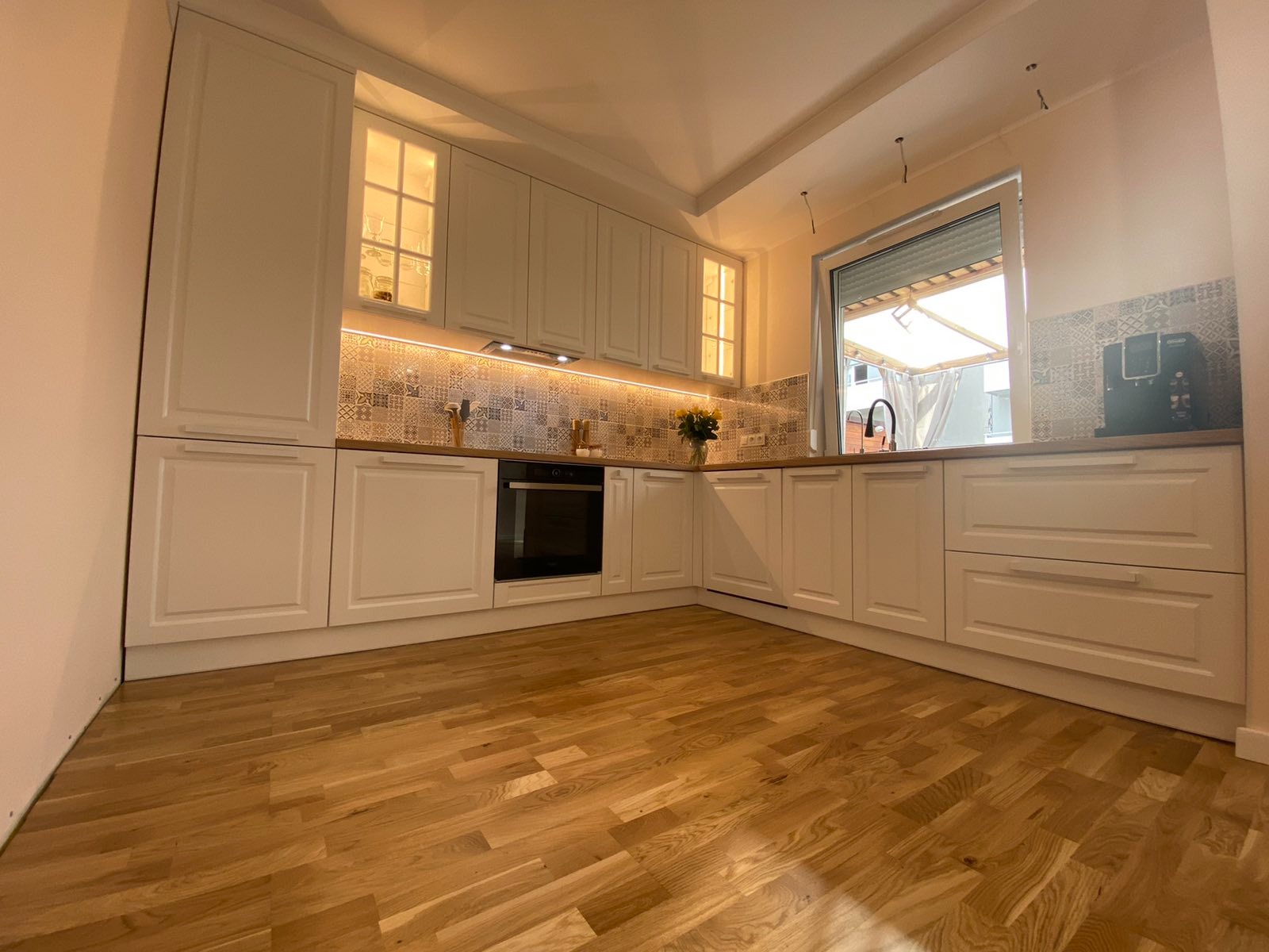 Jasna kuchnia z białymi frontami, drewnianym blatem i podświetleniem LED nad blatem, widok na drewnianą podłogę i okno z roletą zewnętrzną.