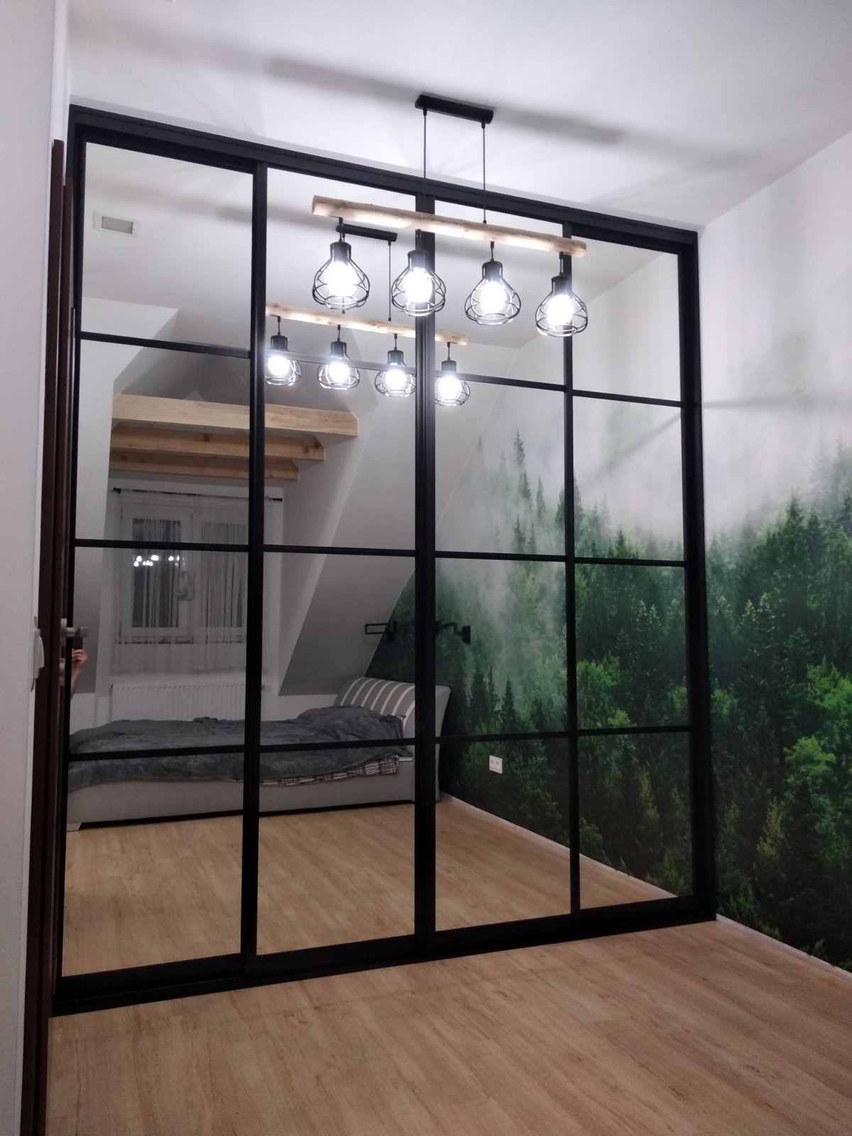 Przeszklona ściana działowa z czarnymi szprosami oddzielająca sypialnię na poddaszu od reszty pomieszczenia, widoczna lampa wisząca z sześcioma żarówkami i fototapeta z motywem lasu.