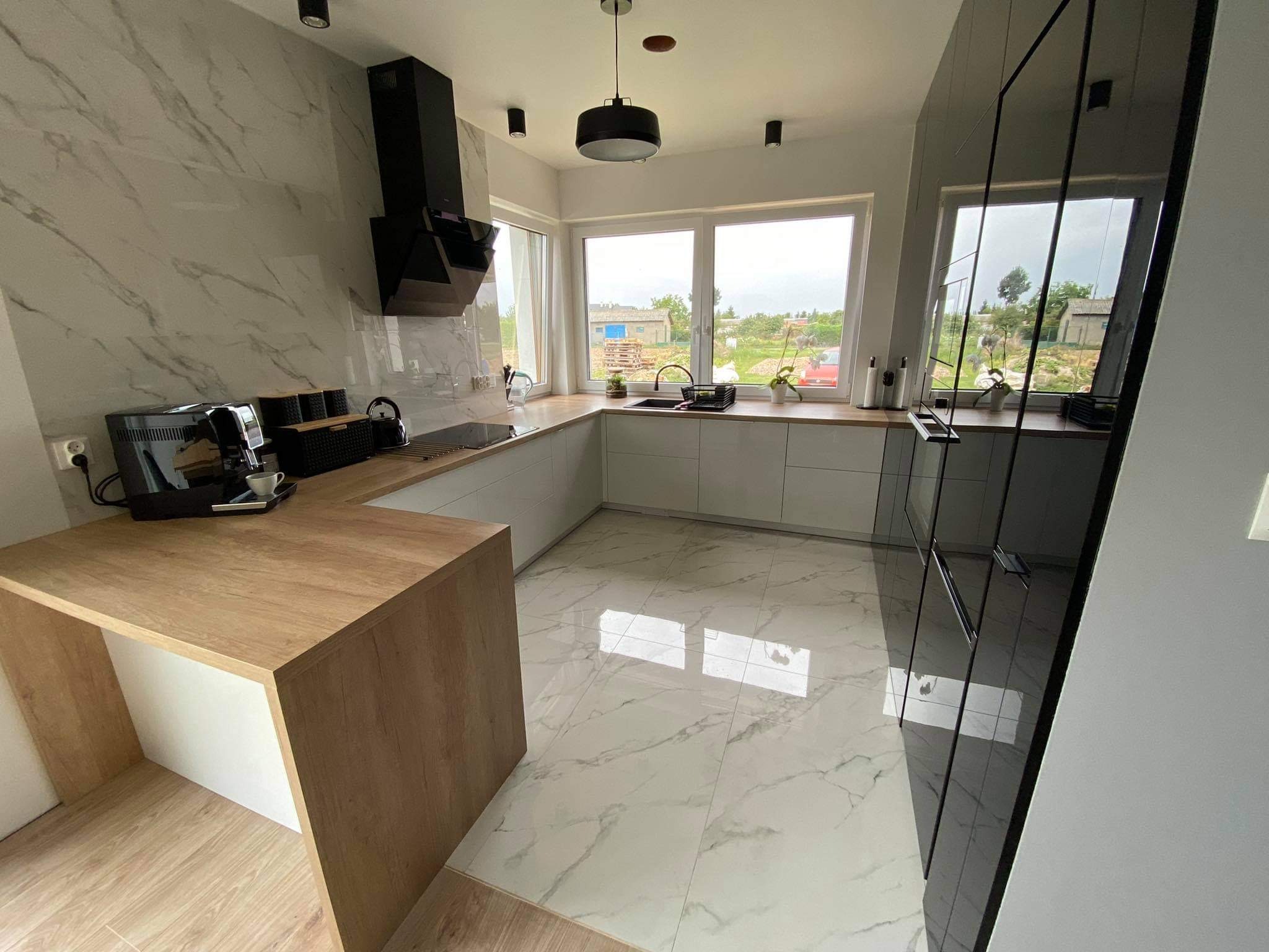Nowoczesna kuchnia z białymi szafkami, drewnianym blatem i marmurową podłogą, widok na ogród przez okno.