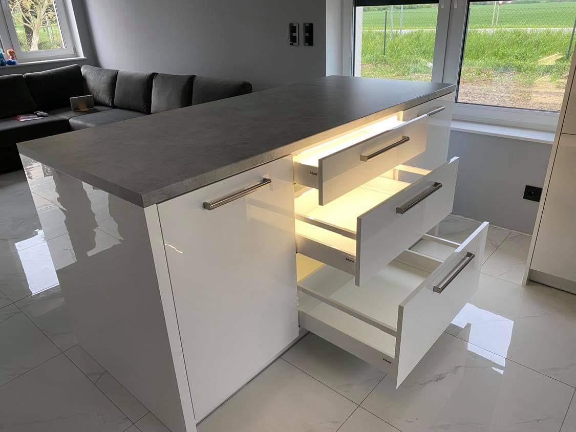 Biała wyspa kuchenna z szarym blatem, otwarte szuflady z wewnętrznym oświetleniem LED, widoczne okucia Blum.