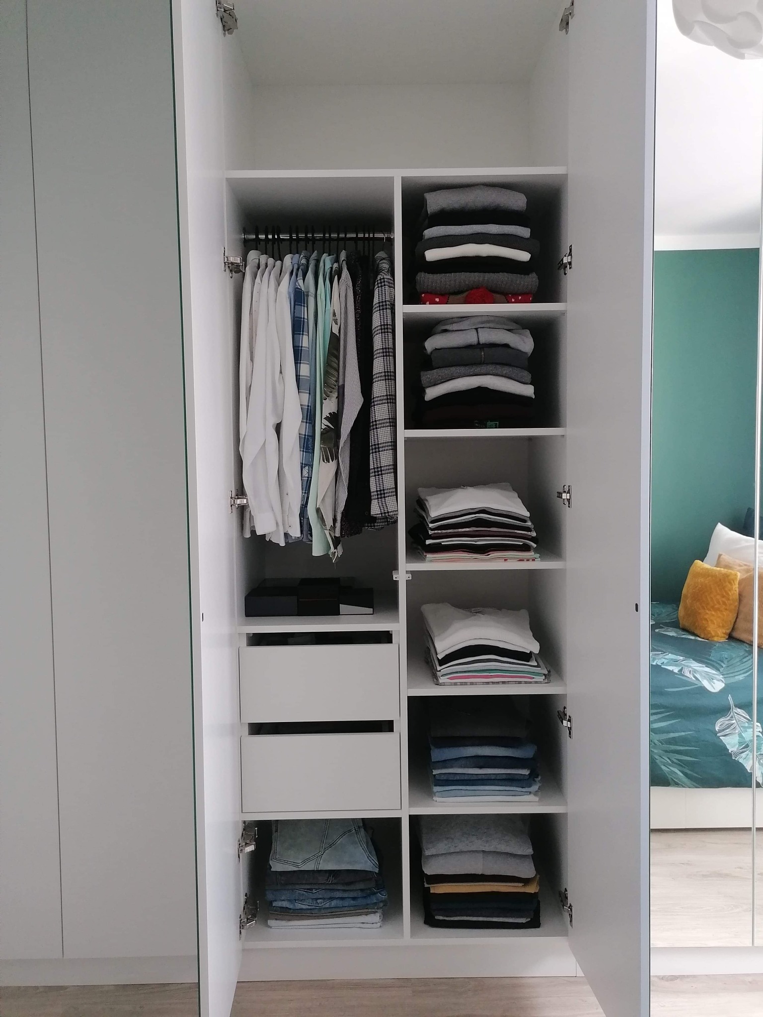 Wnętrze białej szafy na wymiar w Smolcu z drzwiami uchylnymi, widoczne ubrania na wieszakach i półkach, odbicie pokoju w lustrzanych drzwiach.