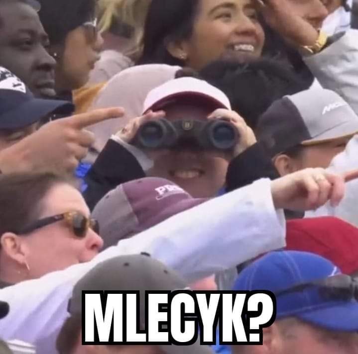 Tłum ludzi, w centrum kobieta z lornetką, a na dole napis 'MLECYK?'