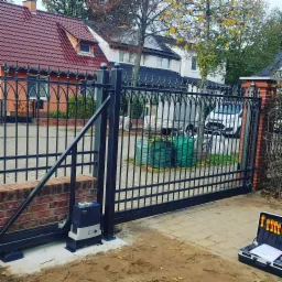 Czarna, metalowa brama wjazdowa z automatycznym napędem, widoczna skrzynka z narzędziami podczas montażu, na tle domy z czerwonymi dachami i jesienne drzewa.