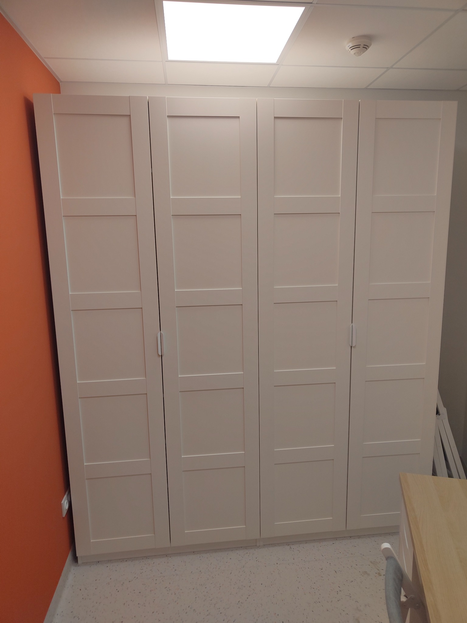 Czterodrzwiowa, biała szafa z panelowymi drzwiami, ustawiona przy pomarańczowej ścianie w pomieszczeniu z podwieszanym sufitem.