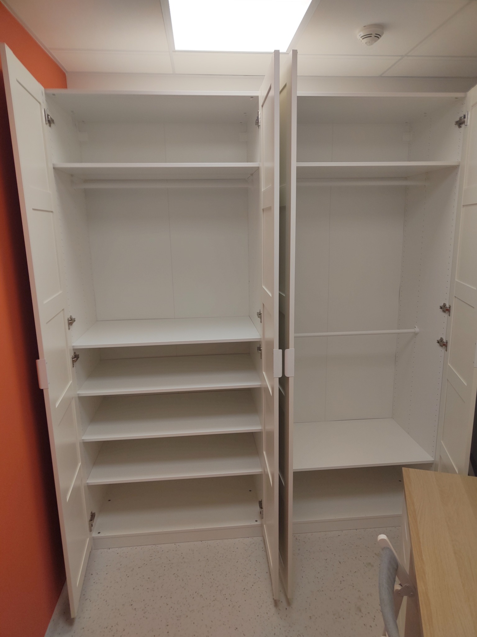 Wnętrze szafy z otwartymi drzwiami, widoczne półki i drążki, białe wykończenie, na podłodze beżowe lastryko, pomarańczowa ściana w tle po lewej stronie.