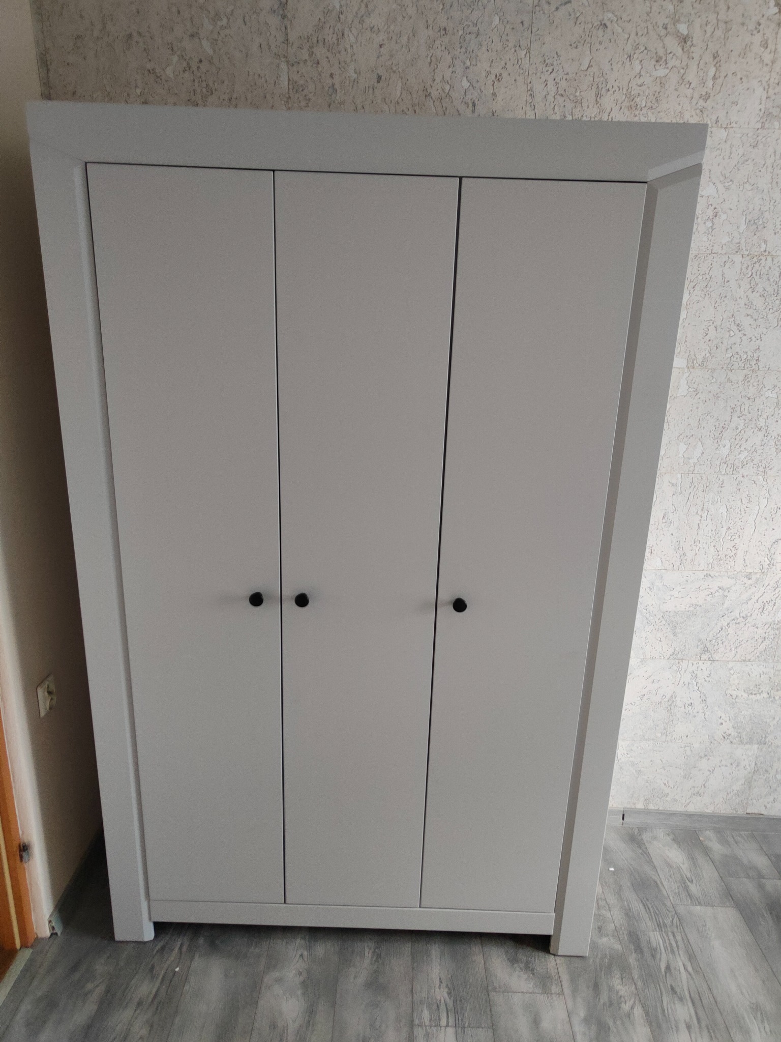 Trzydrzwiowa, szara szafa z okrągłymi, czarnymi uchwytami na tle ściany o nieregularnej fakturze i szarej podłogi imitującej drewno.