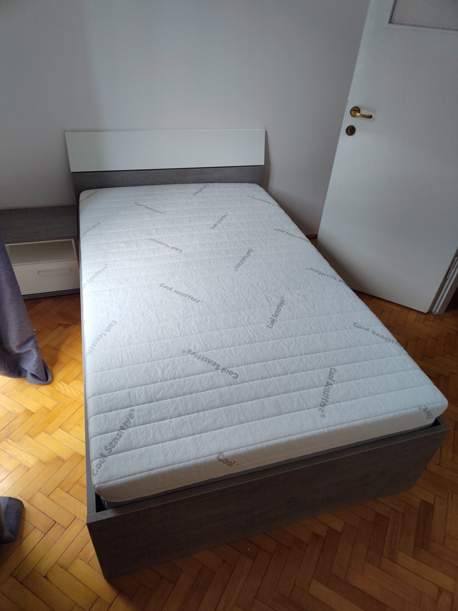 Nowoczesne łóżko jednoosobowe z szarym obramowaniem i białym zagłówkiem, materac z napisem 'Cool Sensitive', obok szafka nocna i białe drzwi, parkiet w jodełkę.