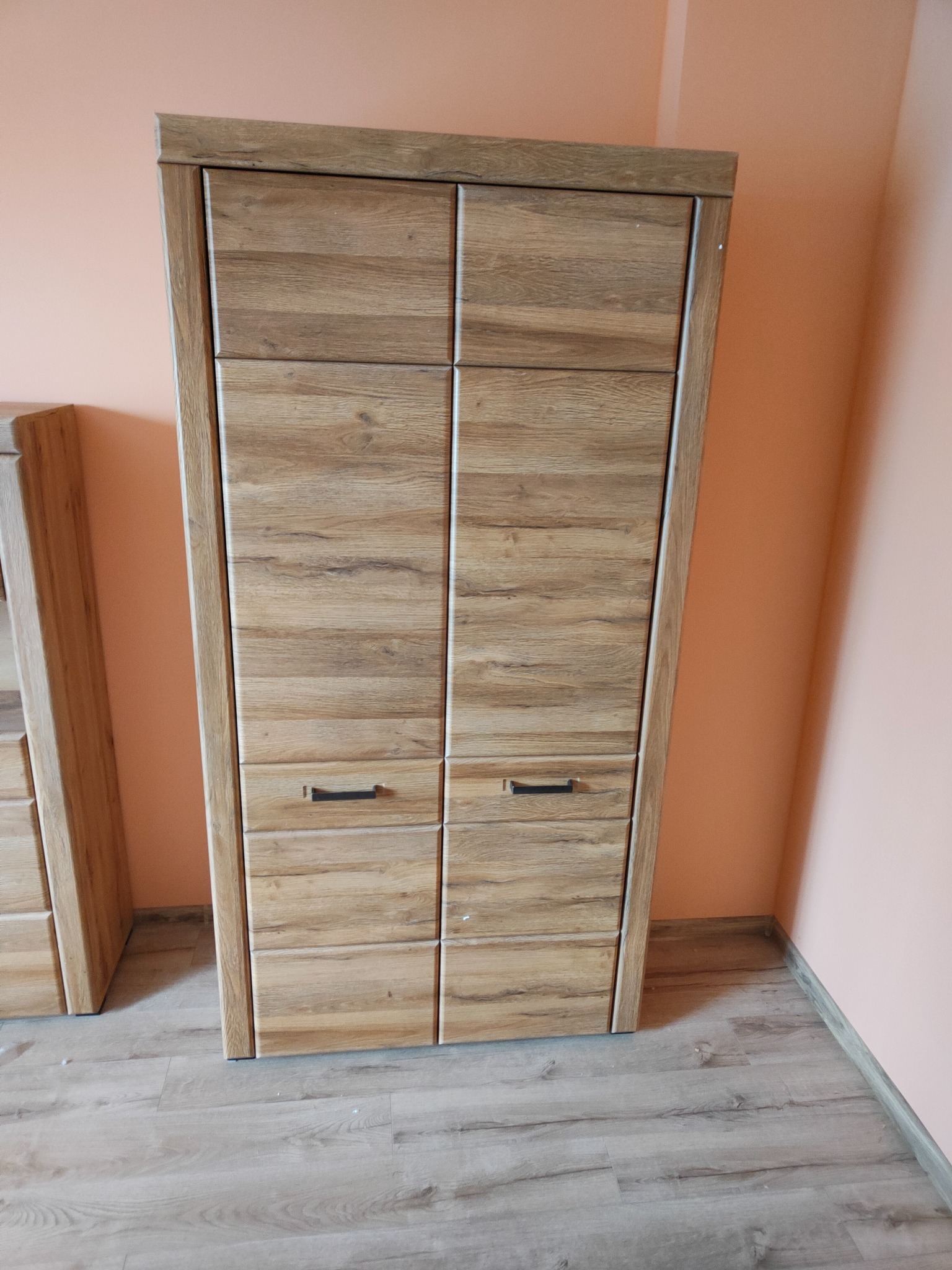 Wysoka, wąska szafa z dwiema parami drzwi i dwiema szufladami, wykończona okleiną imitującą jasne drewno, stoi na drewnianej podłodze przy pomarańczowej ścianie.