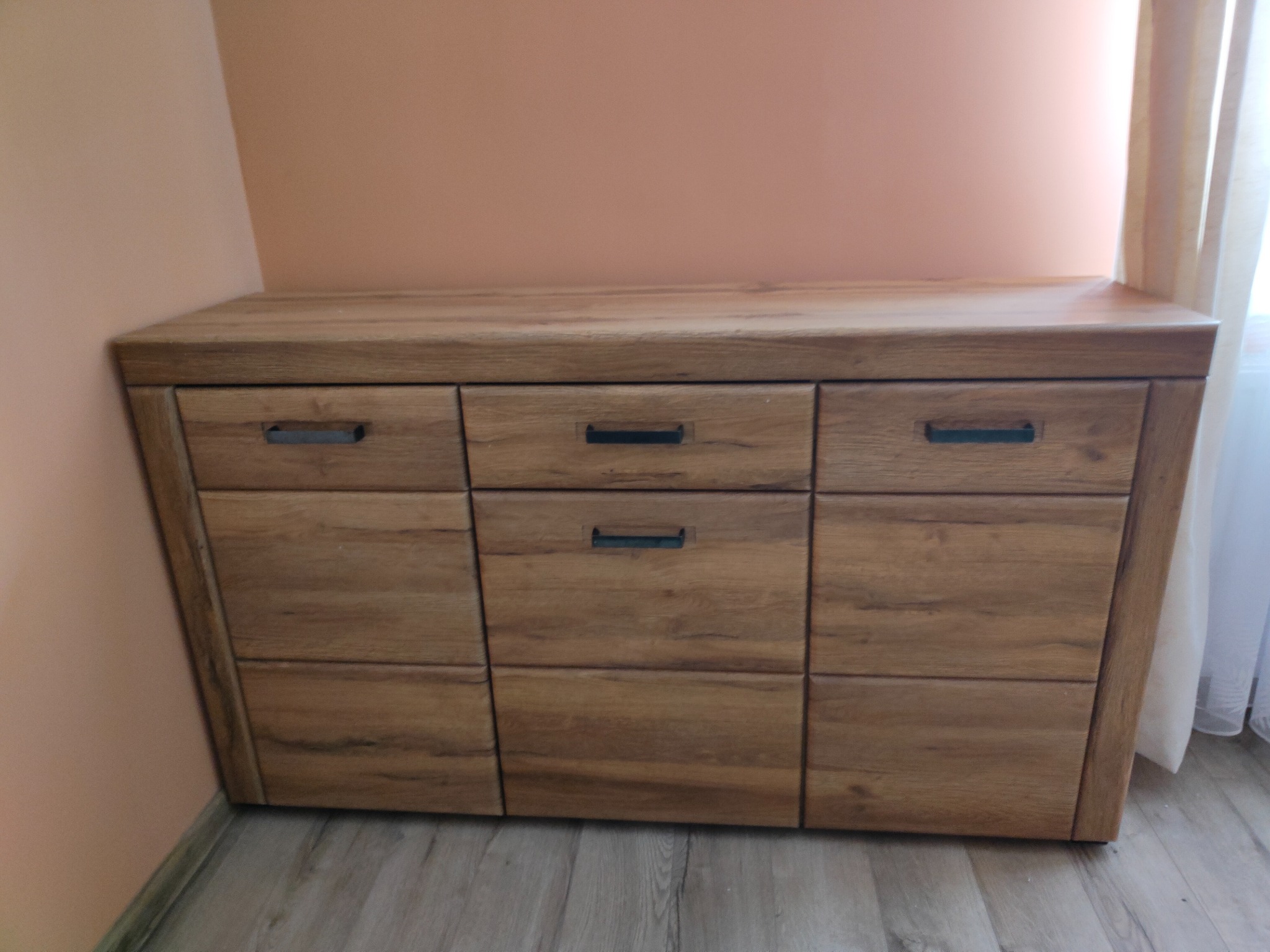 Komoda z sześcioma szufladami w kolorze drewna, z czarnymi uchwytami, stojąca przy ścianie pomalowanej na brzoskwiniowo, z widocznym fragmentem zasłony w tle.