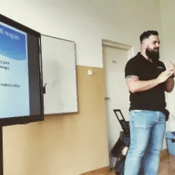 Instruktor z brodą demonstruje gestykulacją podczas szkolenia, obok tablica interaktywna z prezentacją, w tle biała ściana z drzwiami oznaczonymi numerem 123.