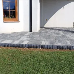 Szara kostka brukowa tworzy taras przy budynku z oknami, obrzeża z palisady. W tle widoczne elementy gabionowe i zieleń.