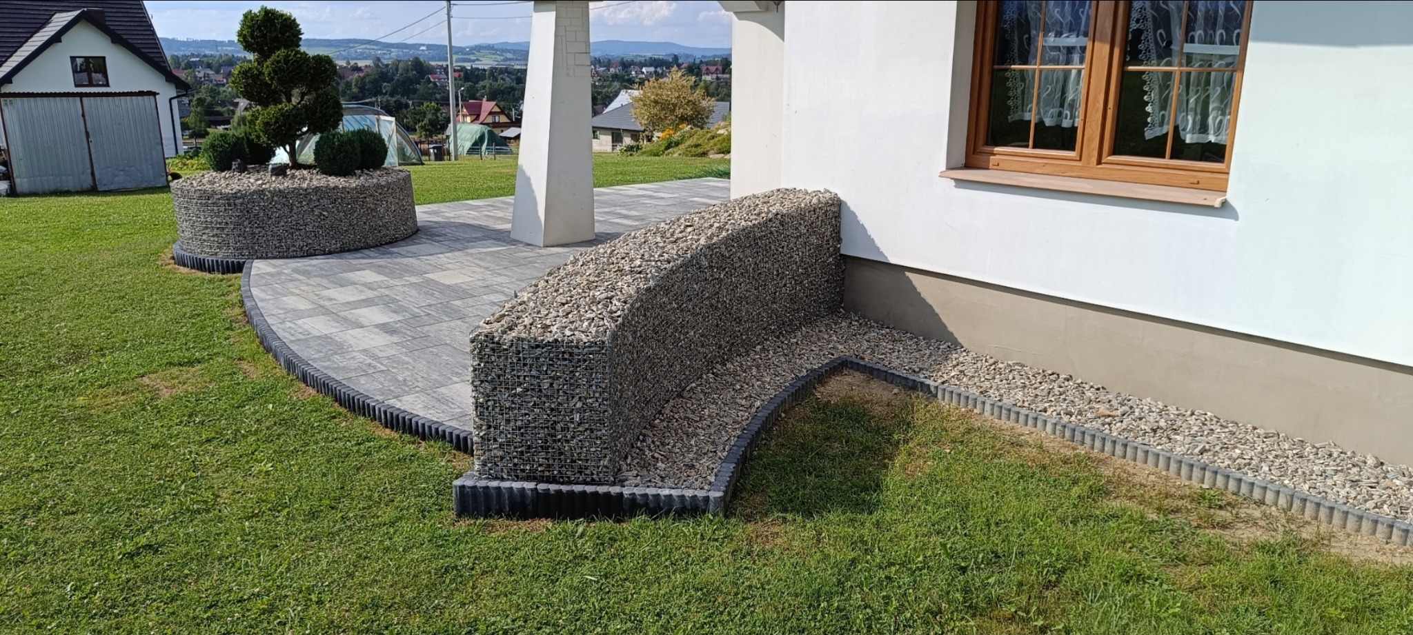 Aranżacja ogrodu z kostki brukowej i gabionów: rabata z drzewkiem bonsai, ścieżka i obrzeże wzdłuż ściany domu. Widok na zielony trawnik i zabudowania w tle.