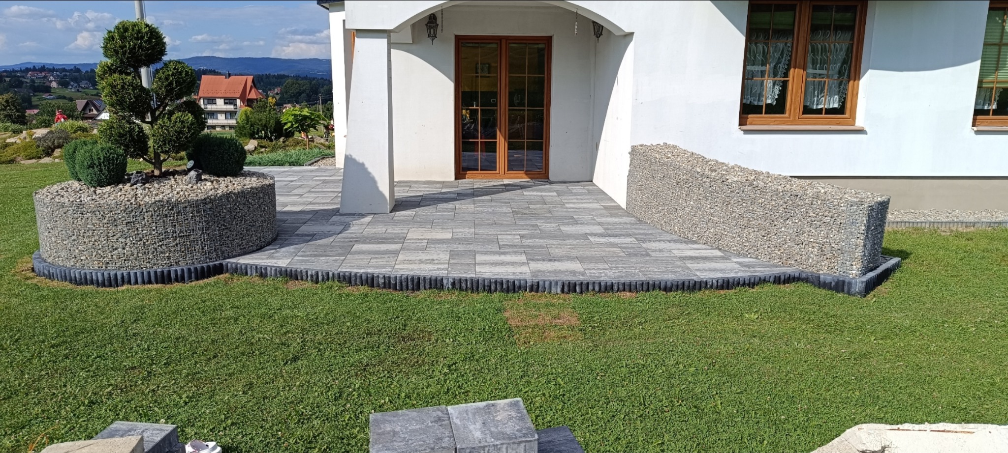 Nowoczesny taras z kostki brukowej w odcieniach szarości, otoczony zielonym trawnikiem. Dekoracyjny element z kamieni w siatce i kształtne drzewko dodają elegancji.