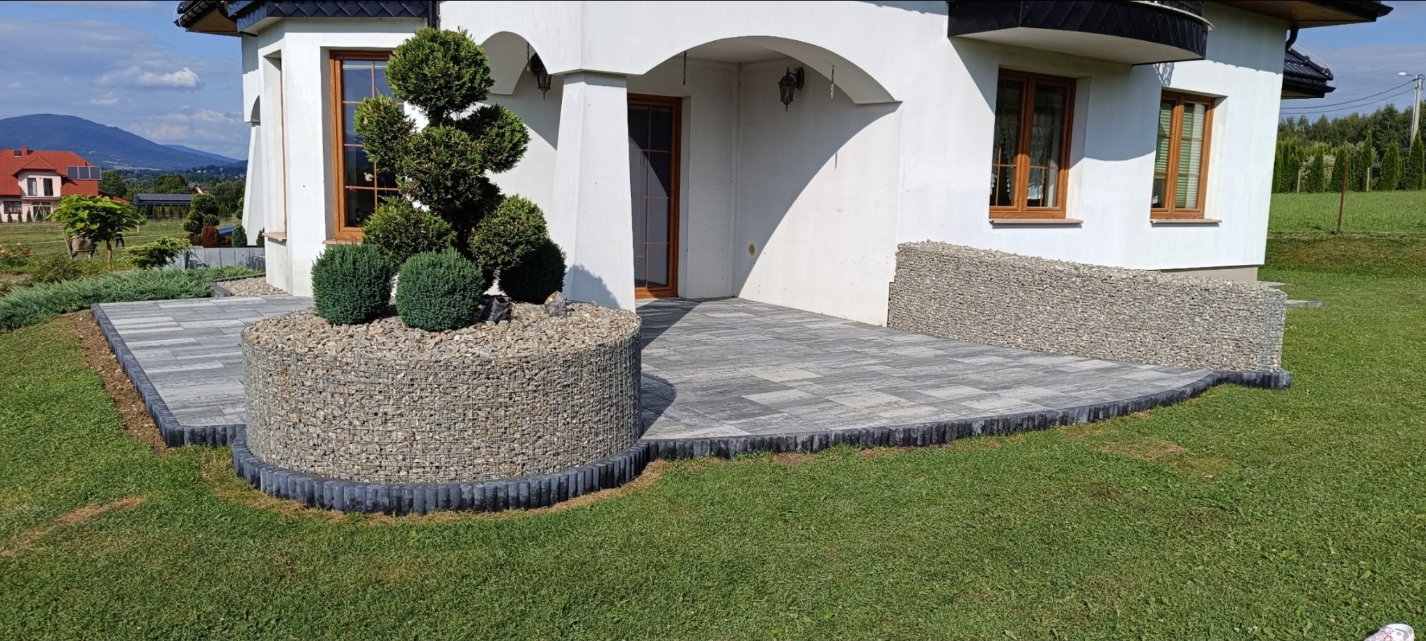 Stylowy taras z kostki brukowej w odcieniach szarości, otoczony gabionami wypełnionymi kamieniami. Na tarasie roślina ozdobna. W tle biały dom z drewnianymi oknami i zielony trawnik.