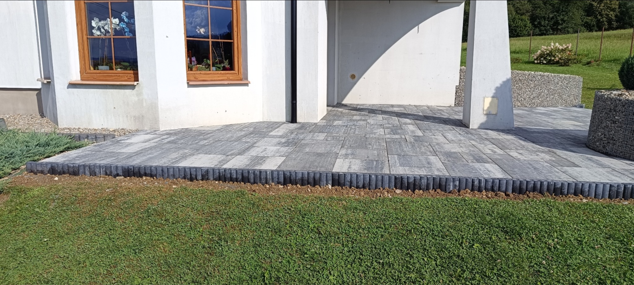 Szara kostka brukowa tworzy taras przy budynku z oknami, obrzeża z palisady. W tle widoczne elementy gabionowe i zieleń.