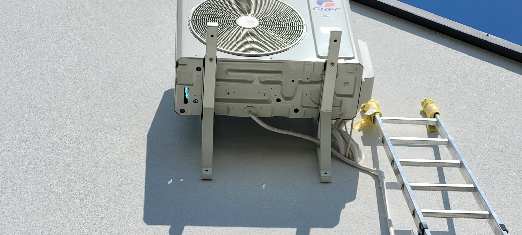Zewnętrzna jednostka klimatyzacji Gree zamontowana na białej elewacji budynku, widoczna drabina z żółtymi osłonami na szczeblach.