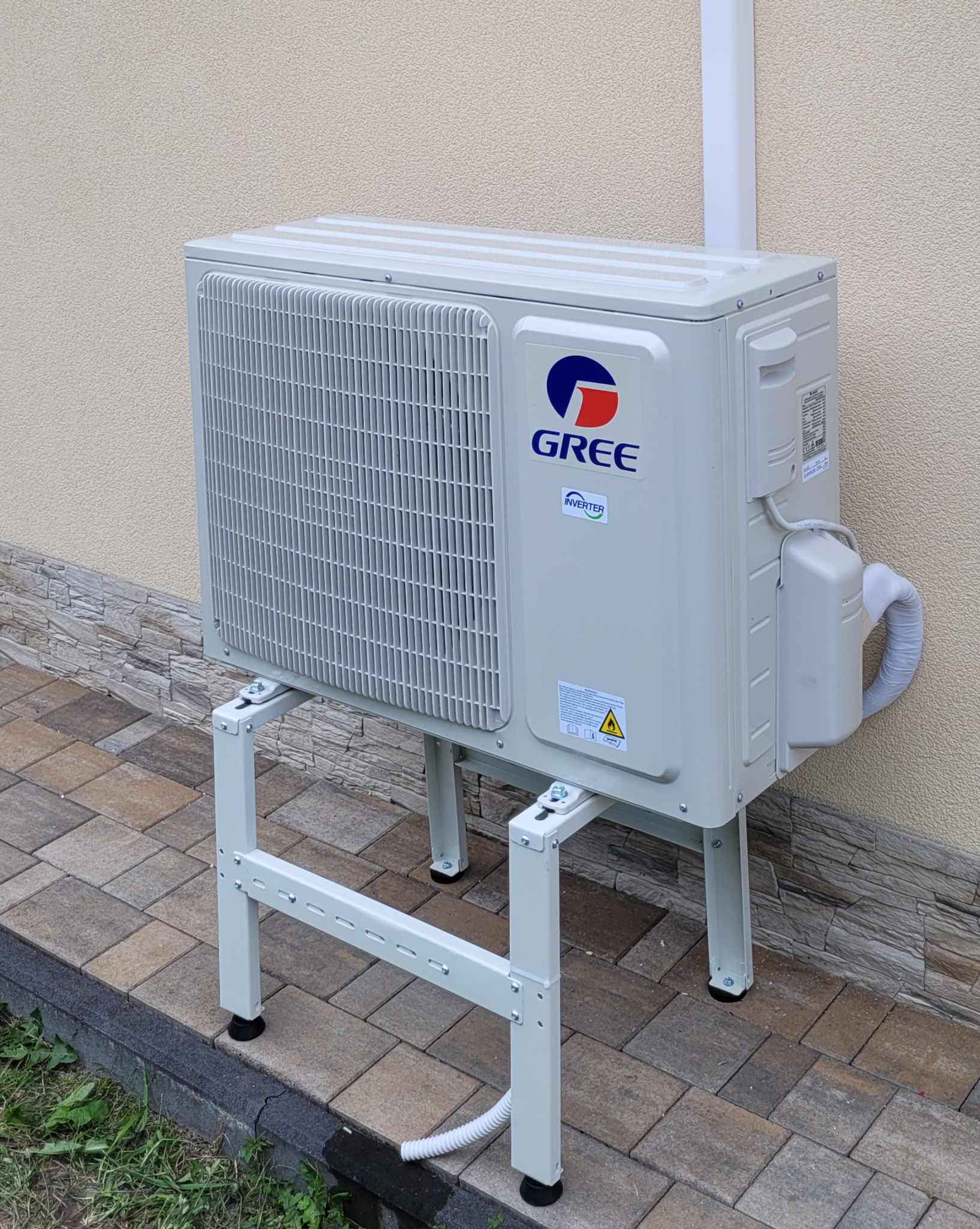 Zewnętrzna jednostka klimatyzacji Gree na metalowym stelażu, zainstalowana przy elewacji budynku z kostki brukowej