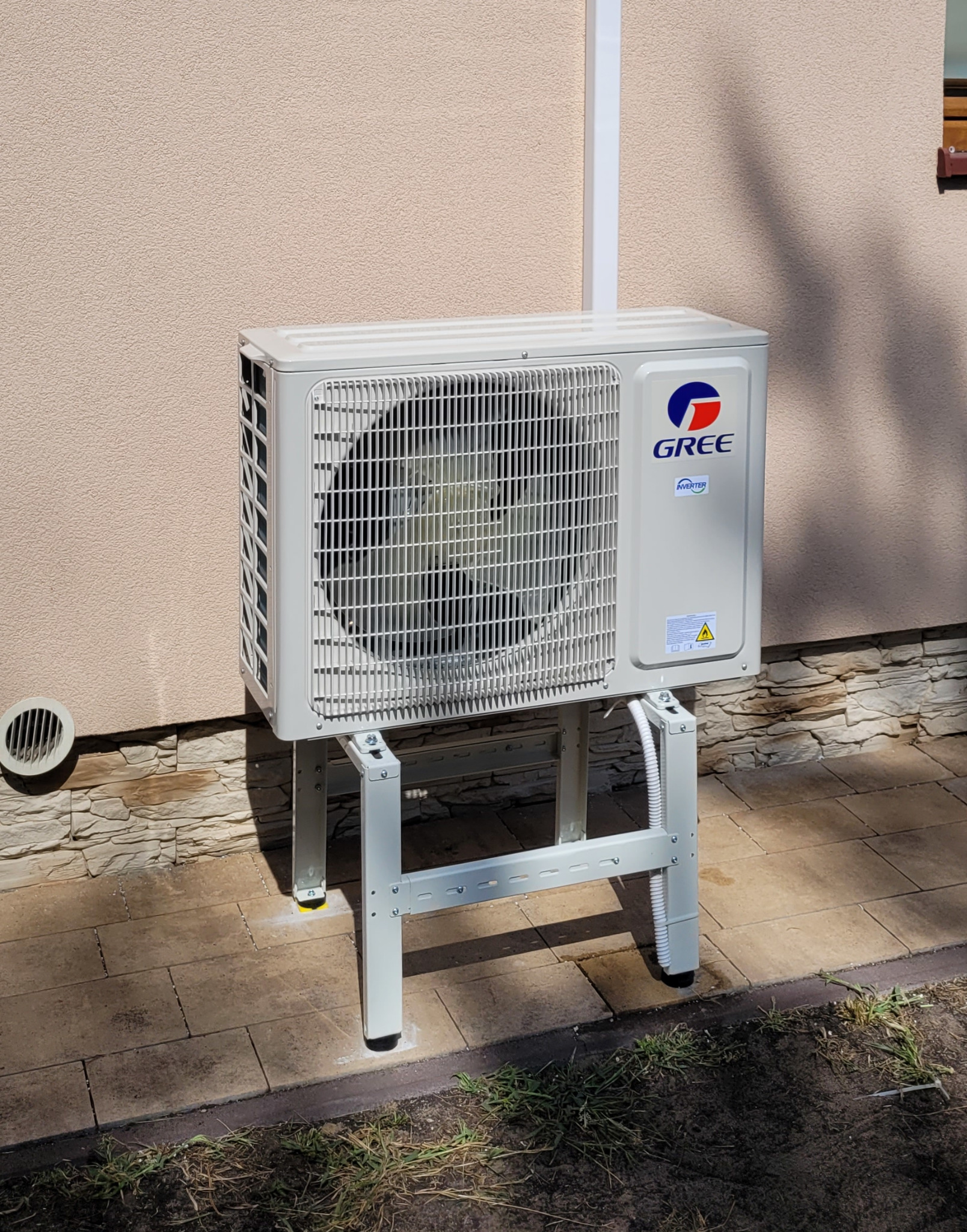 Zewnętrzna jednostka klimatyzacji marki Gree, zamontowana na metalowym stelażu przy ścianie budynku z elementami kamiennej elewacji.
