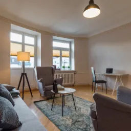 Jasne, przestronne wnętrze gabinetu z dwoma oknami, sofą z poduszkami, fotelami, stolikiem kawowym i biurkiem z laptopem, oświetlone lampą stojącą i wiszącą.