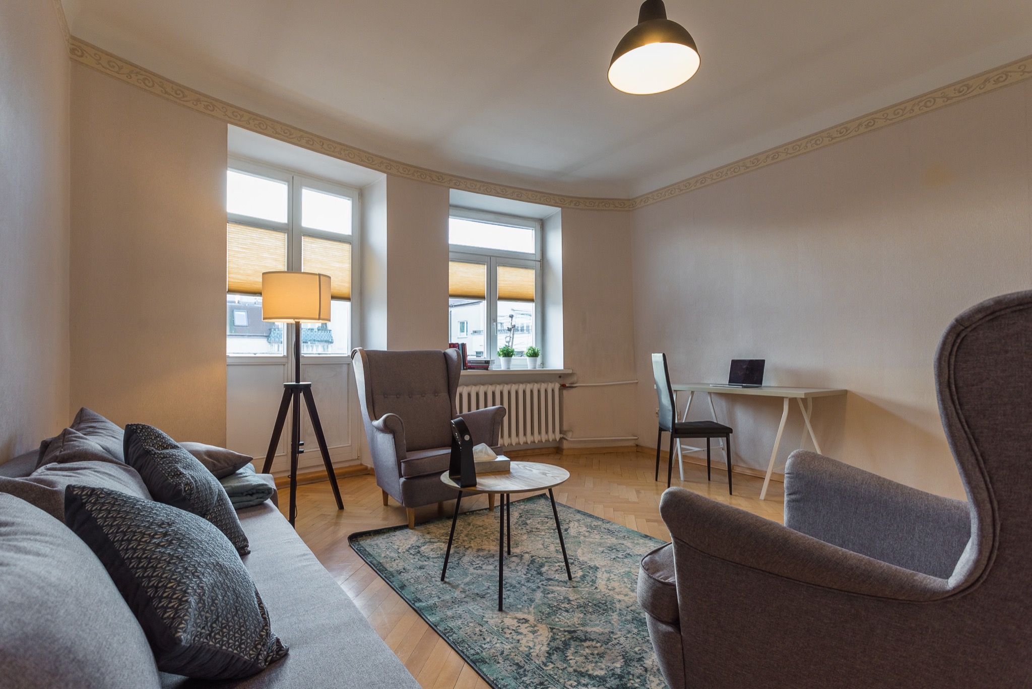 Jasne, przestronne wnętrze gabinetu z dwoma oknami, sofą z poduszkami, fotelami, stolikiem kawowym i biurkiem z laptopem, oświetlone lampą stojącą i wiszącą.