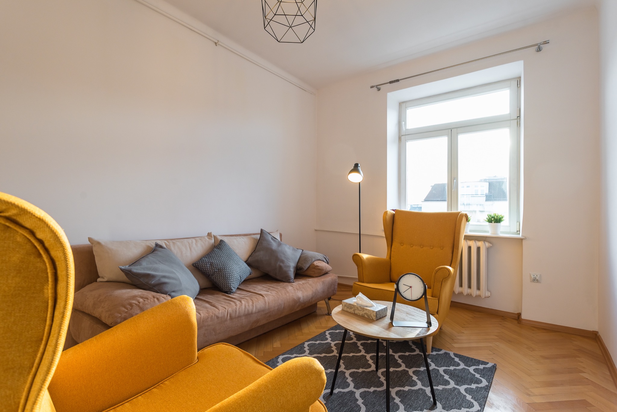 Jasne wnętrze z żółtymi fotelami, brązową kanapą z poduszkami, okrągłym stolikiem z zegarem i pudełkiem chusteczek, na podłodze dywan w marokański wzór, lampa stojąca przy oknie.